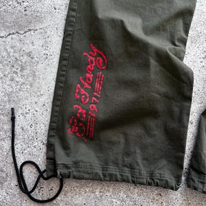May include: Close-up of olive green cargo pants with a drawstring at the hem. The pants feature the red text "Ed Hardy 1971" on the lower leg. The pants are made of a durable fabric.