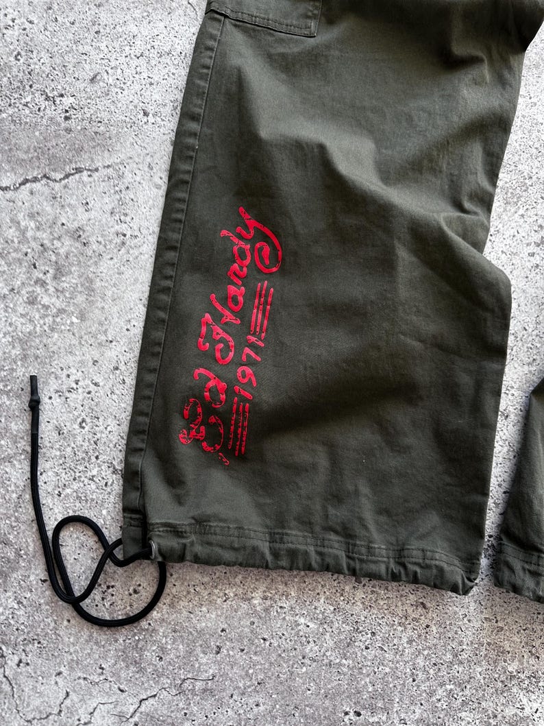 May include: Close-up of olive green cargo pants with a drawstring at the hem. The pants feature the red text "Ed Hardy 1971" on the lower leg. The pants are made of a durable fabric.