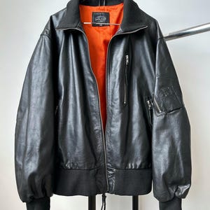 May include: Black leather jacket with a black collar and cuffs, and an orange interior. The jacket has a zipper closure and multiple pockets. The jacket is hanging on a metal rack.