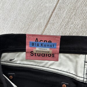 May include: Black denim jeans featuring a pink rectangular brand label on the interior waistband. The label displays the text Acne, Bl&aring; Konst, Floragatan 13, Stockholm, and Studios in black and blue lettering. Copper-colored metal rivets are visible.