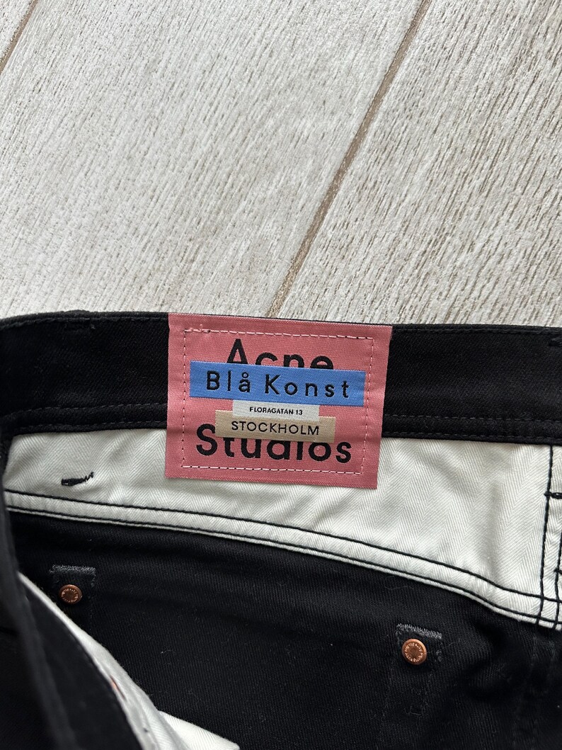 May include: Black denim jeans featuring a pink rectangular brand label on the interior waistband. The label displays the text Acne, Bl&aring; Konst, Floragatan 13, Stockholm, and Studios in black and blue lettering. Copper-colored metal rivets are visible.