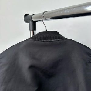 May include: A black bomber jacket featuring a smooth, satin-like finish and a ribbed knit collar. The jacket is suspended from a metal hanger on a clothing rack, showcasing the upper back and neckline area of the garment in a neutral setting.