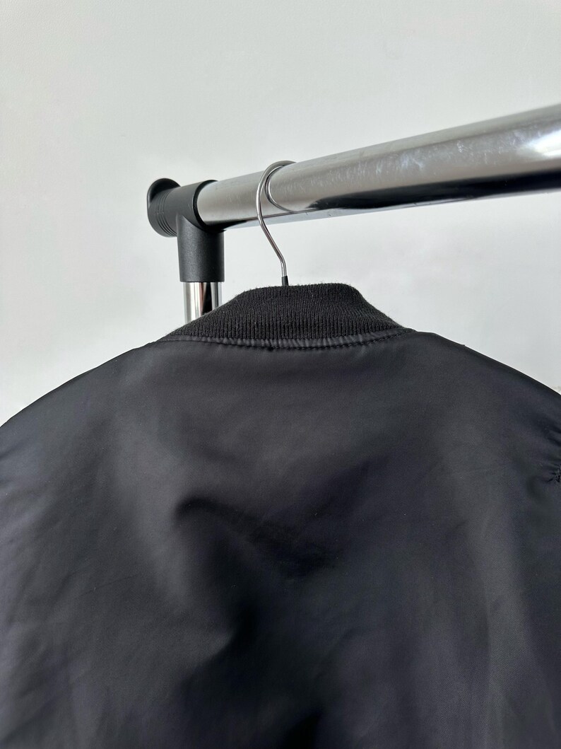 May include: A black bomber jacket featuring a smooth, satin-like finish and a ribbed knit collar. The jacket is suspended from a metal hanger on a clothing rack, showcasing the upper back and neckline area of the garment in a neutral setting.