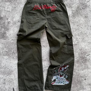May include: Olive green cargo pants with a drawstring waist and multiple pockets. The back features red embroidered text, and the lower leg has a colorful embroidered design of a koi fish and waves.