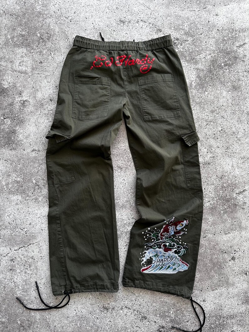 May include: Olive green cargo pants with a drawstring waist and multiple pockets. The back features red embroidered text, and the lower leg has a colorful embroidered design of a koi fish and waves.
