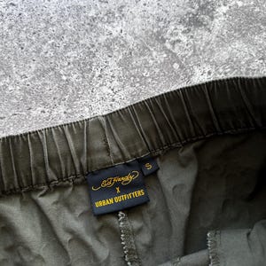 May include: Close-up of olive green drawstring pants with a visible elastic waistband. A black tag with gold lettering reads "Ed Hardy x Urban Outfitters" and the size is "S". The pants are made of a soft, textured fabric.