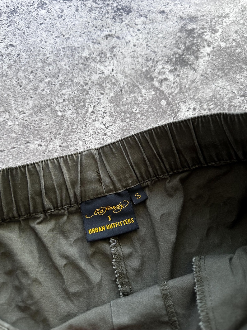 May include: Close-up of olive green drawstring pants with a visible elastic waistband. A black tag with gold lettering reads "Ed Hardy x Urban Outfitters" and the size is "S". The pants are made of a soft, textured fabric.