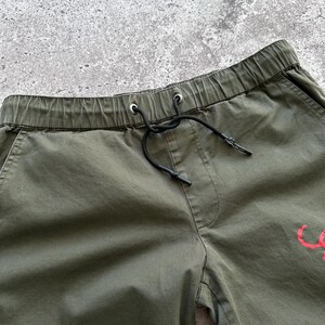 May include: Close-up of olive green drawstring shorts. The shorts have an elastic waistband with a black drawstring. A small red graphic is visible on the shorts. The shorts are made of a lightweight fabric.