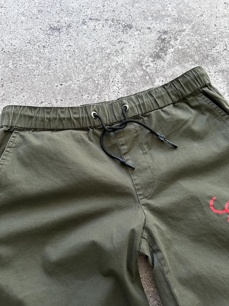 May include: Close-up of olive green drawstring shorts. The shorts have an elastic waistband with a black drawstring. A small red graphic is visible on the shorts. The shorts are made of a lightweight fabric.