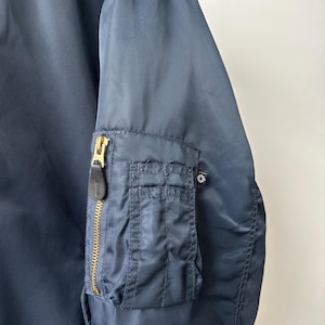 May include: Navy blue bomber jacket with a gold zipper and a small pocket on the left sleeve. The jacket has a classic design and is made of a smooth, durable material. The jacket is a versatile piece of outerwear.