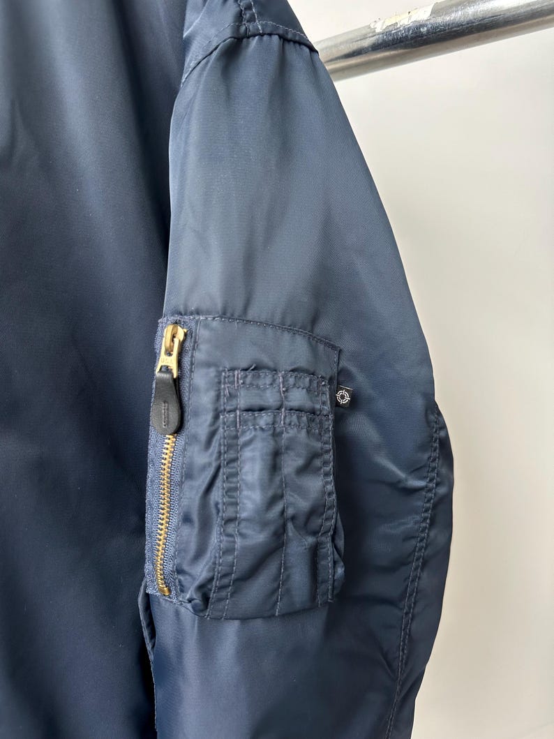 May include: Navy blue bomber jacket with a gold zipper and a small pocket on the left sleeve. The jacket has a classic design and is made of a smooth, durable material. The jacket is a versatile piece of outerwear.