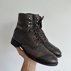 May include: A pair of dark brown leather lace-up boots featuring a textured finish, a rounded toe cap, and silver-toned metal eyelets. The boots have a mid-calf height with dark brown laces and a solid black sole, showcasing a structured design.