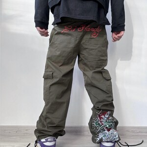 May include: Olive green cargo pants with the text "Ed Hardy" in red across the waistband. The pants feature a decorative design on the lower leg and are paired with purple and white sneakers.