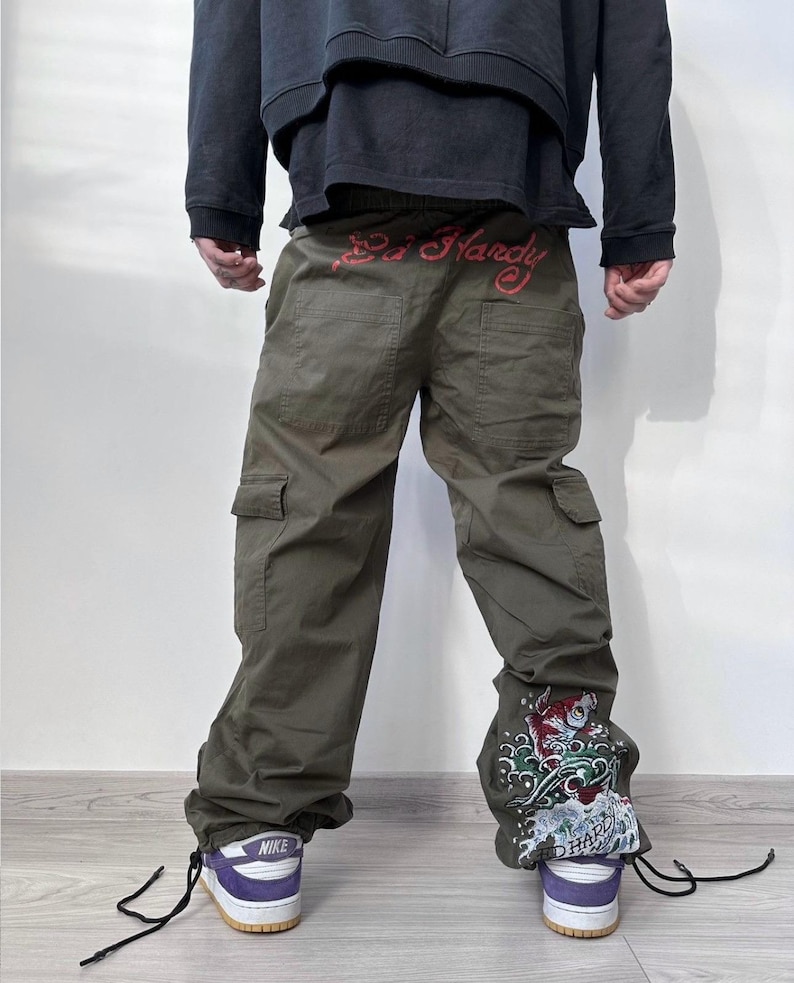 May include: Olive green cargo pants with the text "Ed Hardy" in red across the waistband. The pants feature a decorative design on the lower leg and are paired with purple and white sneakers.
