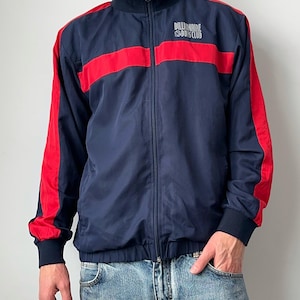 May include: Navy blue track jacket with red stripes across the chest and down the sleeves. The jacket has a full zipper and a stand-up collar. The text "BILLIONAIRE BOYS CLUB" is printed on the chest in gray.