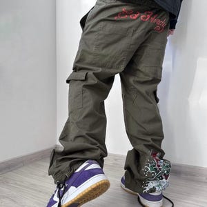 May include: Olive green cargo pants with multiple pockets and a drawstring hem. The back of the pants features red text. The person is wearing purple and white sneakers with a brown sole. The right pant leg has a colorful graphic.