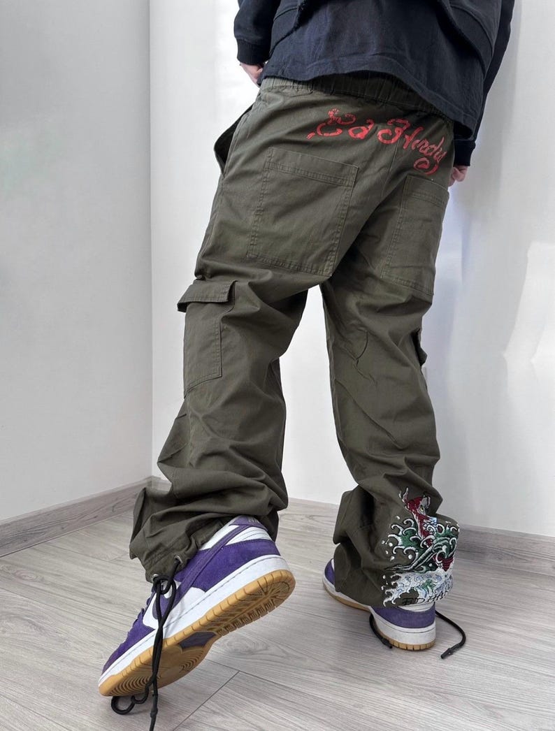 May include: Olive green cargo pants with multiple pockets and a drawstring hem. The back of the pants features red text. The person is wearing purple and white sneakers with a brown sole. The right pant leg has a colorful graphic.