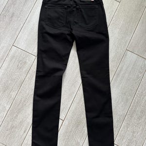 May include: A pair of solid black denim pants featuring a classic five-pocket design, a waistband with belt loops, and a rectangular black leather-like brand patch positioned on the back right side of the waistband above the rear pocket.