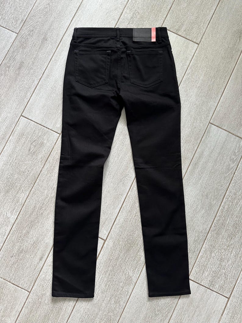 May include: A pair of solid black denim pants featuring a classic five-pocket design, a waistband with belt loops, and a rectangular black leather-like brand patch positioned on the back right side of the waistband above the rear pocket.