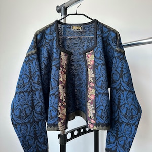 Pode incluir: Cardigã azul e preto estampado com um design floral. O cardigã tem uma borda preta e uma faixa floral na frente. A etiqueta diz "OLEANA".