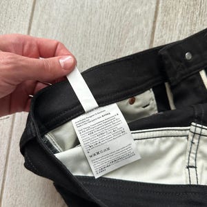 May include: Black denim pants featuring a white interior fabric lining and a prominent white care and content label attached to the inner waistband. The label displays multi-language text including brand information, material composition, and washing instructions.