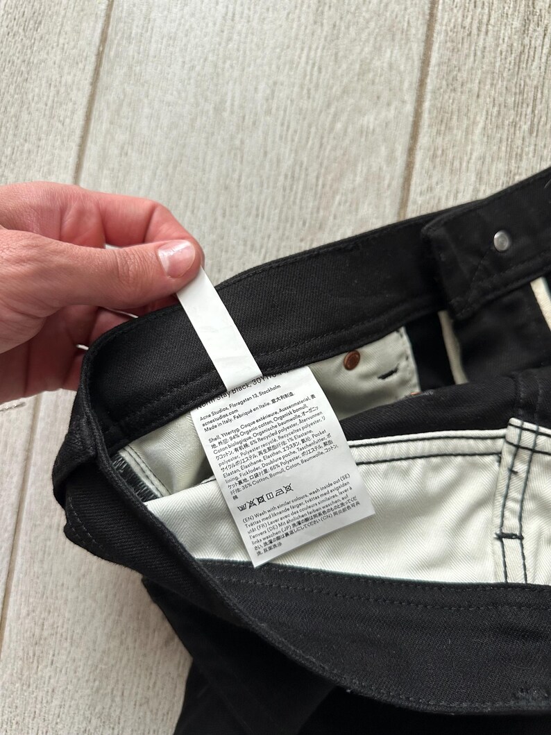 May include: Black denim pants featuring a white interior fabric lining and a prominent white care and content label attached to the inner waistband. The label displays multi-language text including brand information, material composition, and washing instructions.