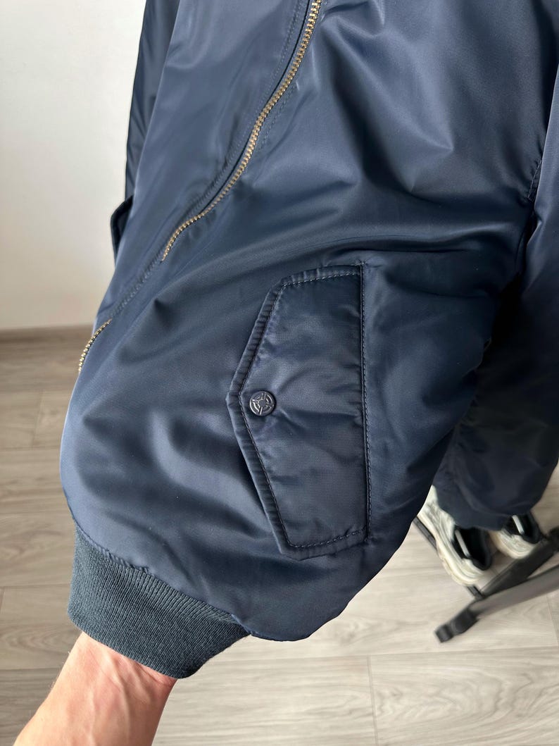May include: Navy blue bomber jacket with a ribbed cuff and a zippered front. The jacket features a pocket with a button closure and a small logo. The jacket is made of a smooth, shiny material.