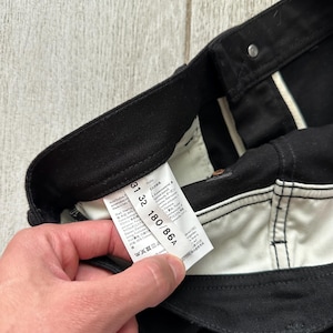 May include: Black trousers featuring a white interior waistband and pocket lining. A white care and size label is attached to the inner waistband, displaying the text Acne Studios, Made in Italy, 31 32, and 180/86A, along with various care symbols.