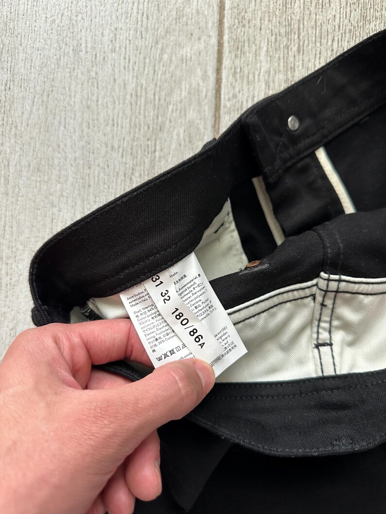May include: Black trousers featuring a white interior waistband and pocket lining. A white care and size label is attached to the inner waistband, displaying the text Acne Studios, Made in Italy, 31 32, and 180/86A, along with various care symbols.