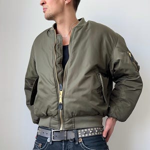 Peut inclure: Veste de bombardier vert olive avec col et poignets côtelés. La veste a une fermeture éclair dorée et une petite poche sur la manche gauche. La personne porte un débardeur noir, un jean délavé foncé et une ceinture cloutée.