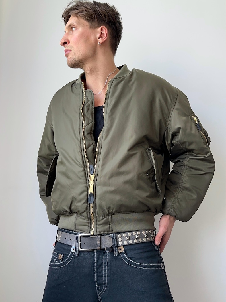 May include: Olive green bomber jacket with a ribbed collar and cuffs. The jacket has a gold zipper and a small pocket on the left sleeve. The person is wearing a black tank top, dark wash jeans, and a studded belt.