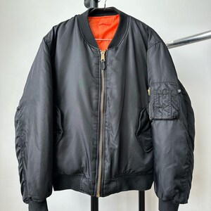 May include: A black bomber jacket featuring a bright orange interior lining, a central gold-toned metal zipper, ribbed black cuffs and hem, and a small utility pocket with a zipper closure located on the upper left sleeve of the garment.