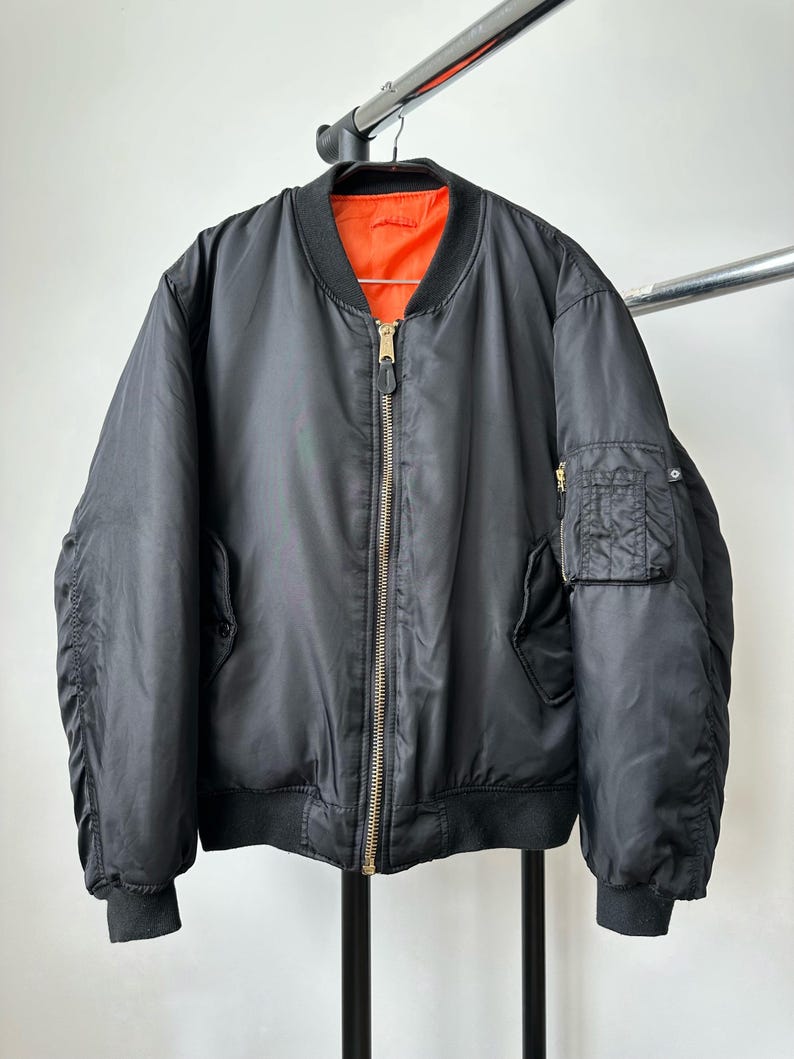 May include: A black bomber jacket featuring a bright orange interior lining, a central gold-toned metal zipper, ribbed black cuffs and hem, and a small utility pocket with a zipper closure located on the upper left sleeve of the garment.