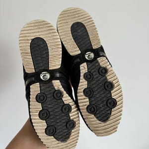 May include: A pair of black Adidas sneakers, viewed from the soles. The soles are cream-colored with a ridged design and black circular accents. The Adidas logo is visible on the shoe's arch. The shoes are held up against a white background.