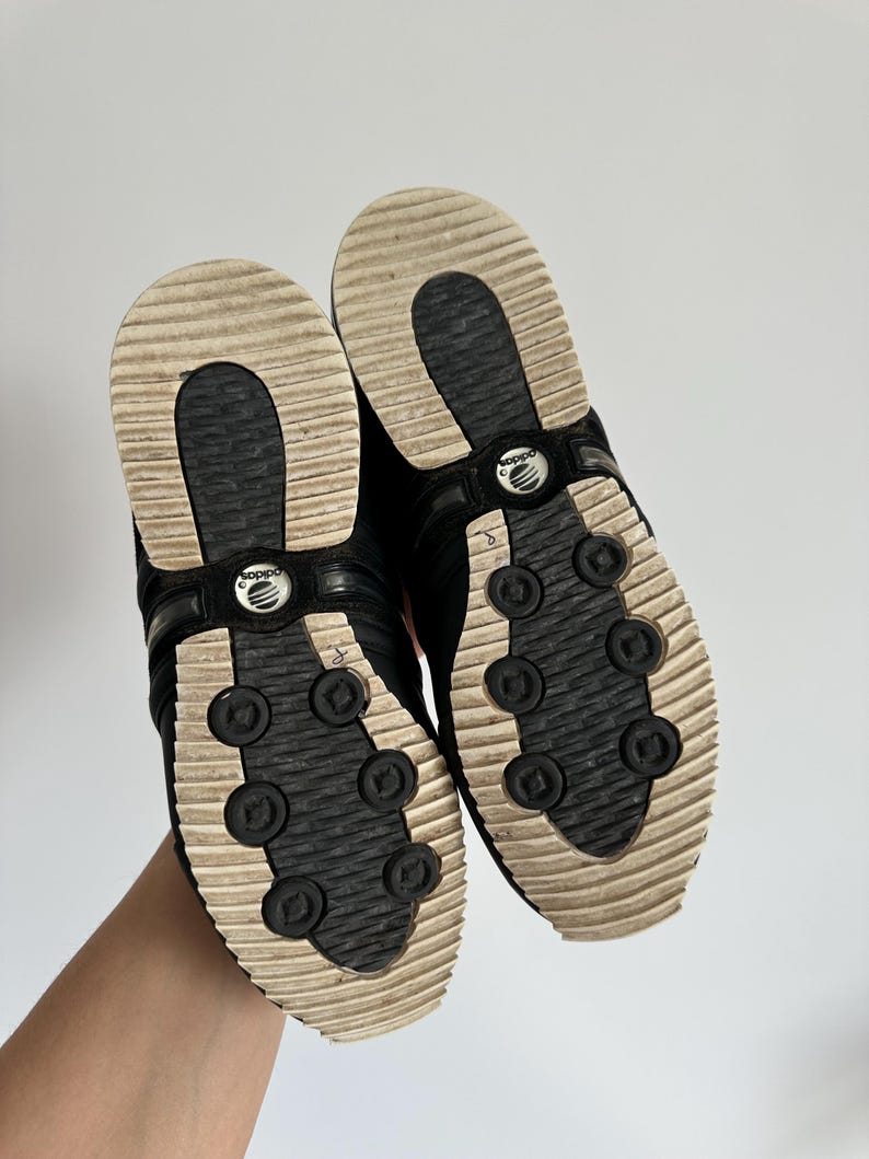 May include: A pair of black Adidas sneakers, viewed from the soles. The soles are cream-colored with a ridged design and black circular accents. The Adidas logo is visible on the shoe's arch. The shoes are held up against a white background.