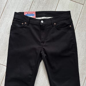 May include: Black denim jeans featuring a classic five-pocket design with a button and zipper fly closure. A pink interior label at the waistband displays the text Acne Bl&aring; Konst in blue lettering, along with additional white text details.