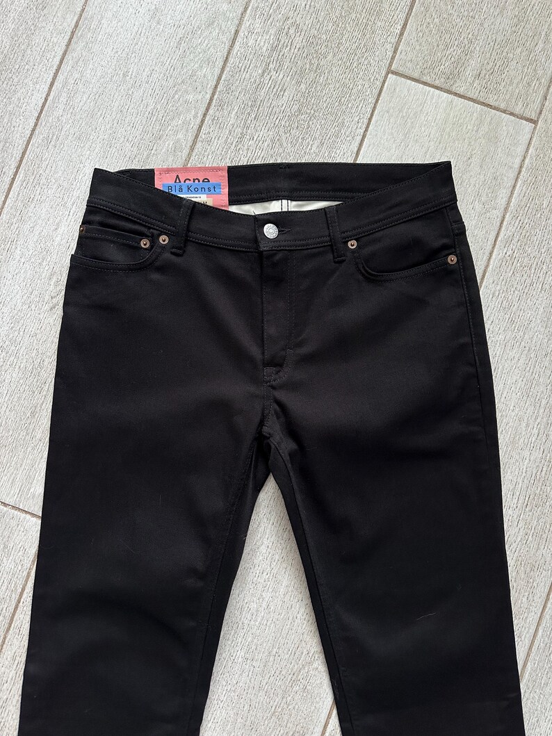 May include: Black denim jeans featuring a classic five-pocket design with a button and zipper fly closure. A pink interior label at the waistband displays the text Acne Bl&aring; Konst in blue lettering, along with additional white text details.