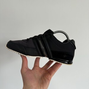 May include: Black athletic shoe with a white sole. The shoe features a black upper with three stripes and a textured design. The shoe is held up by a hand.