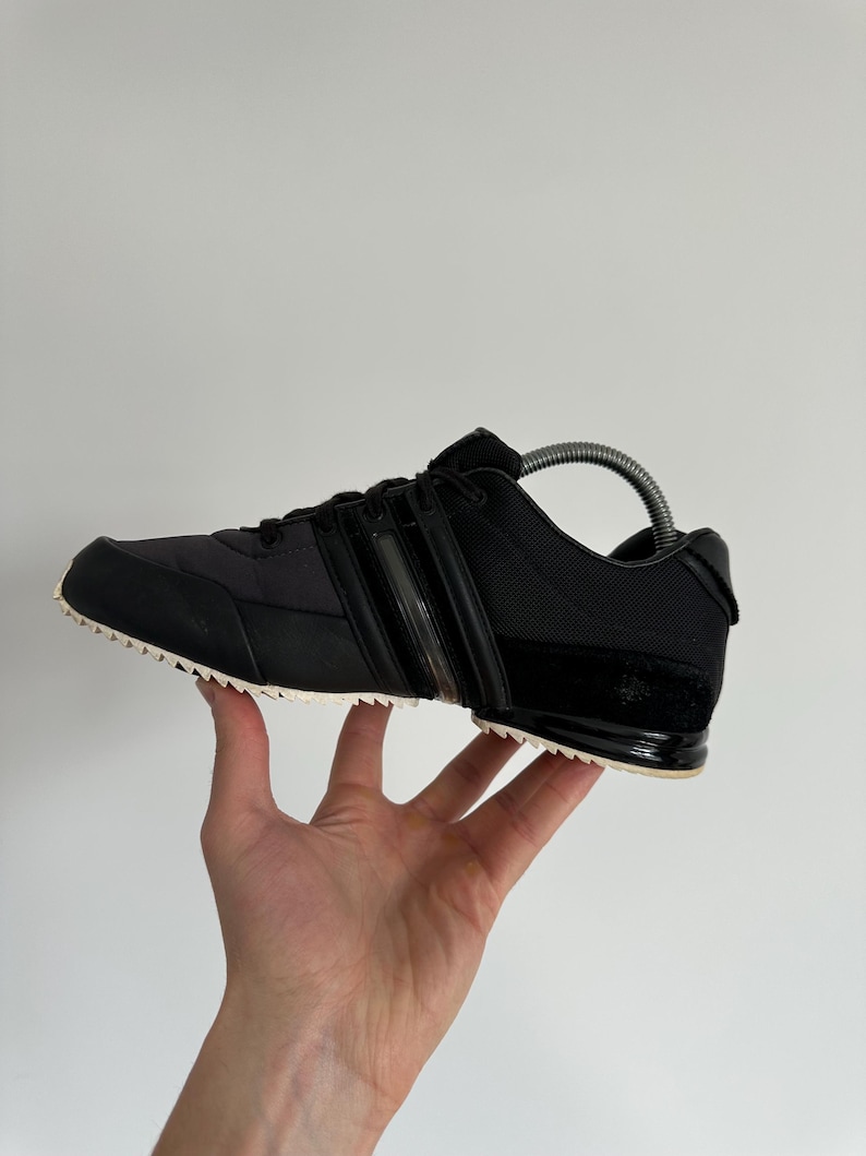 May include: Black athletic shoe with a white sole. The shoe features a black upper with three stripes and a textured design. The shoe is held up by a hand.