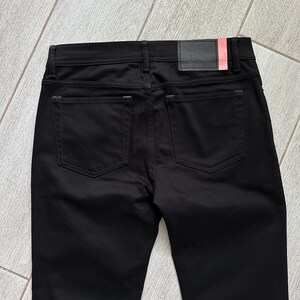 May include: A pair of solid black pants featuring a classic five-pocket design with two back pockets, a waistband with belt loops, and a rectangular black leather-like patch on the back right side with a small vertical red accent stripe.