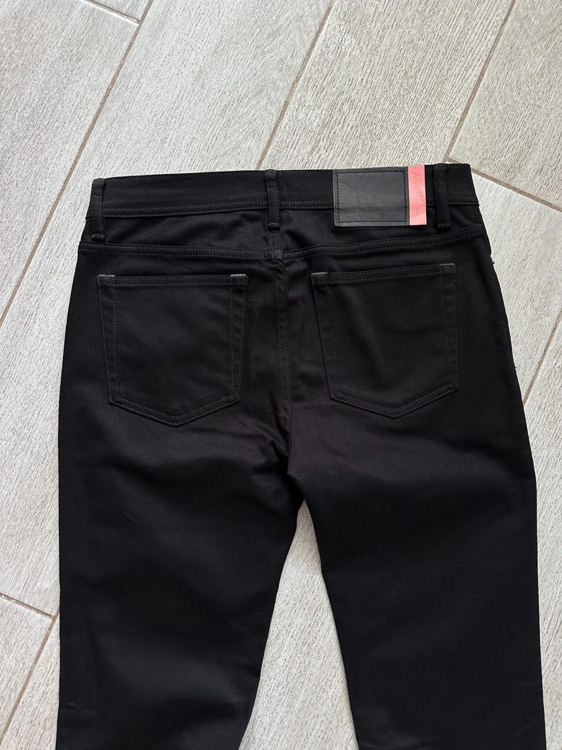 May include: A pair of solid black pants featuring a classic five-pocket design with two back pockets, a waistband with belt loops, and a rectangular black leather-like patch on the back right side with a small vertical red accent stripe.