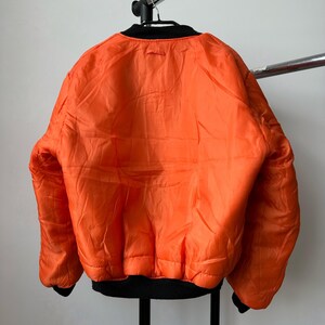 May include: A bright orange bomber jacket featuring a black ribbed collar, black ribbed cuffs, and a black ribbed hem. The jacket has a smooth, quilted-style texture across the back and long sleeves, hanging on a black hanger against a plain wall.