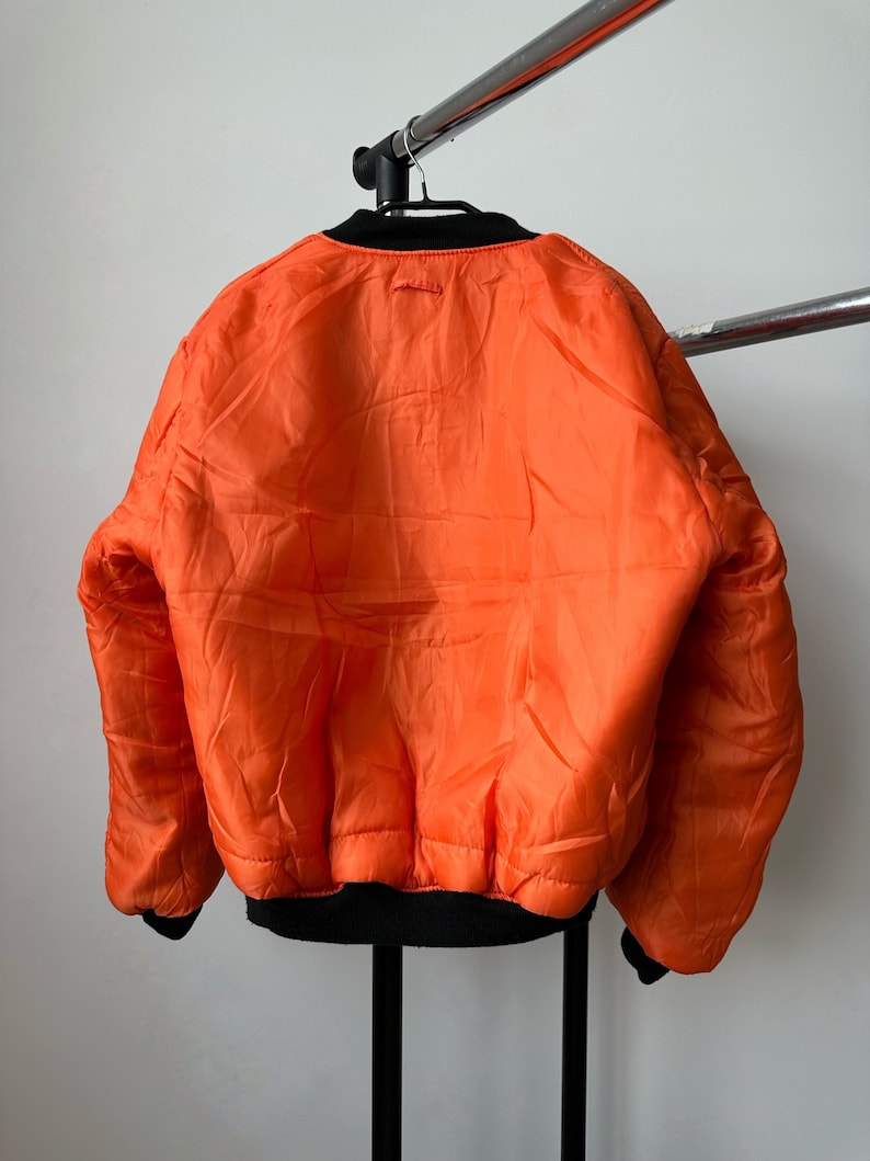 May include: A bright orange bomber jacket featuring a black ribbed collar, black ribbed cuffs, and a black ribbed hem. The jacket has a smooth, quilted-style texture across the back and long sleeves, hanging on a black hanger against a plain wall.
