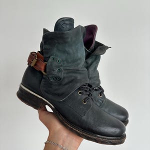 May include: A dark charcoal grey leather ankle boot featuring a slouchy, folded collar design, black laces, and a contrasting brown leather strap with a brass buckle detail. The boot has a rounded toe, a low heel, and a dark purple interior lining.