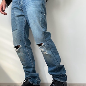 May include: Blue denim jeans featuring a distressed finish with frayed knee holes and scattered silver-tone metal studs throughout the legs. The jeans have a classic five-pocket construction, a button fly, and a straight-leg fit with a medium wash.