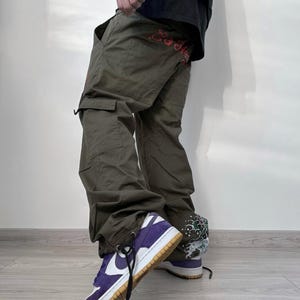 May include: Olive green cargo pants with multiple pockets and red embroidered text. The pants are paired with purple and white sneakers with a brown sole and black laces. The sneakers have a white Nike swoosh.