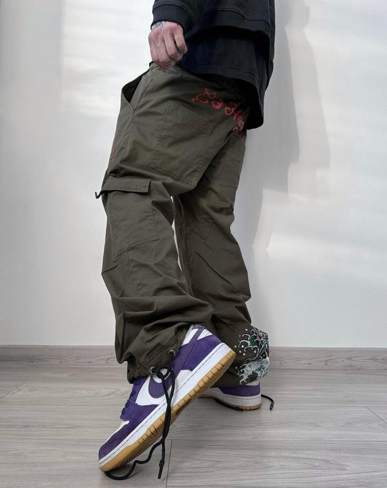 May include: Olive green cargo pants with multiple pockets and red embroidered text. The pants are paired with purple and white sneakers with a brown sole and black laces. The sneakers have a white Nike swoosh.