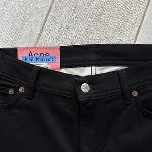 May include: Black denim jeans featuring a pink interior waistband label with the text Acne Bl&aring; Konst, a silver metal button closure, and copper-toned rivets at the pocket corners. The garment has a classic five-pocket construction with visible stitching.