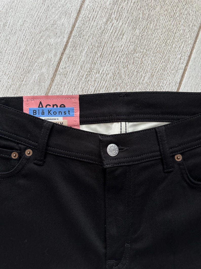 May include: Black denim jeans featuring a pink interior waistband label with the text Acne Bl&aring; Konst, a silver metal button closure, and copper-toned rivets at the pocket corners. The garment has a classic five-pocket construction with visible stitching.