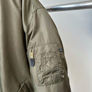 May include: Olive green bomber jacket with a gold zipper and a small pocket on the sleeve. The jacket has a quilted texture and a black zipper pull. A small logo is visible on the sleeve.